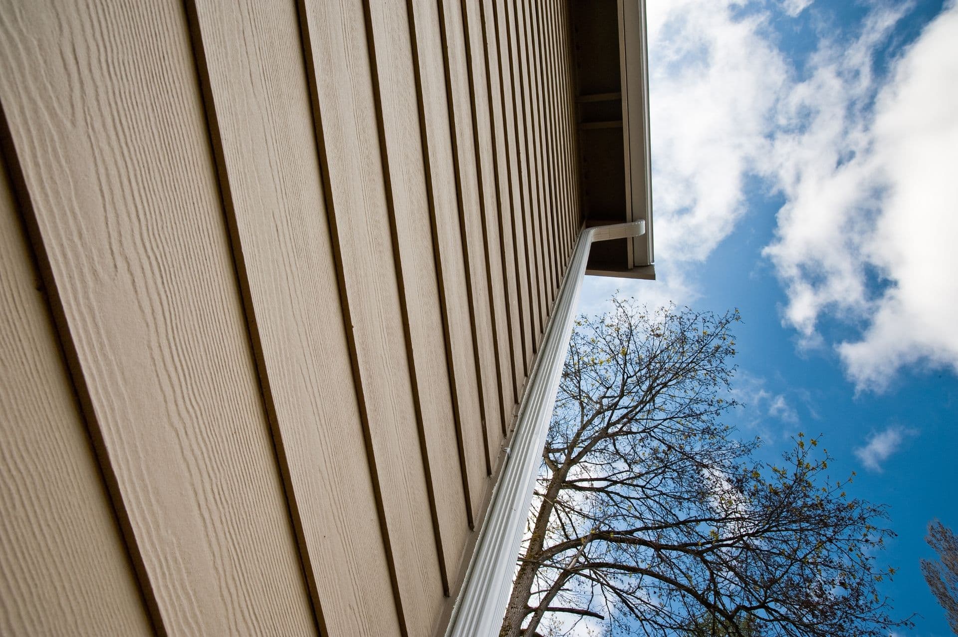 An upward-looking view of a house with new tan siding and white aluminum gutters and downspout.