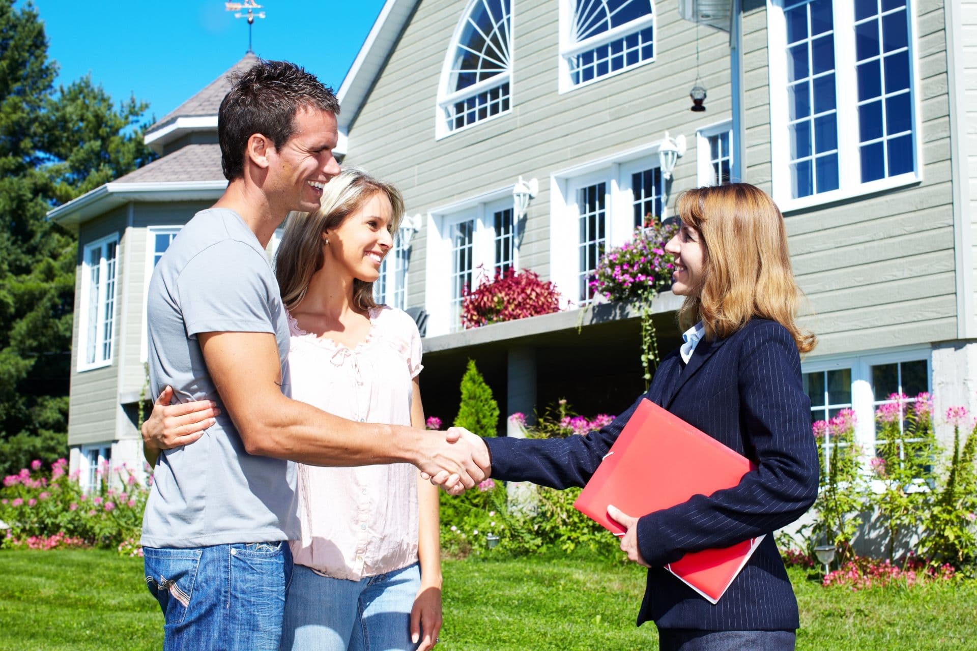 A couple smiles and shakes hands with a professional in front of their house.