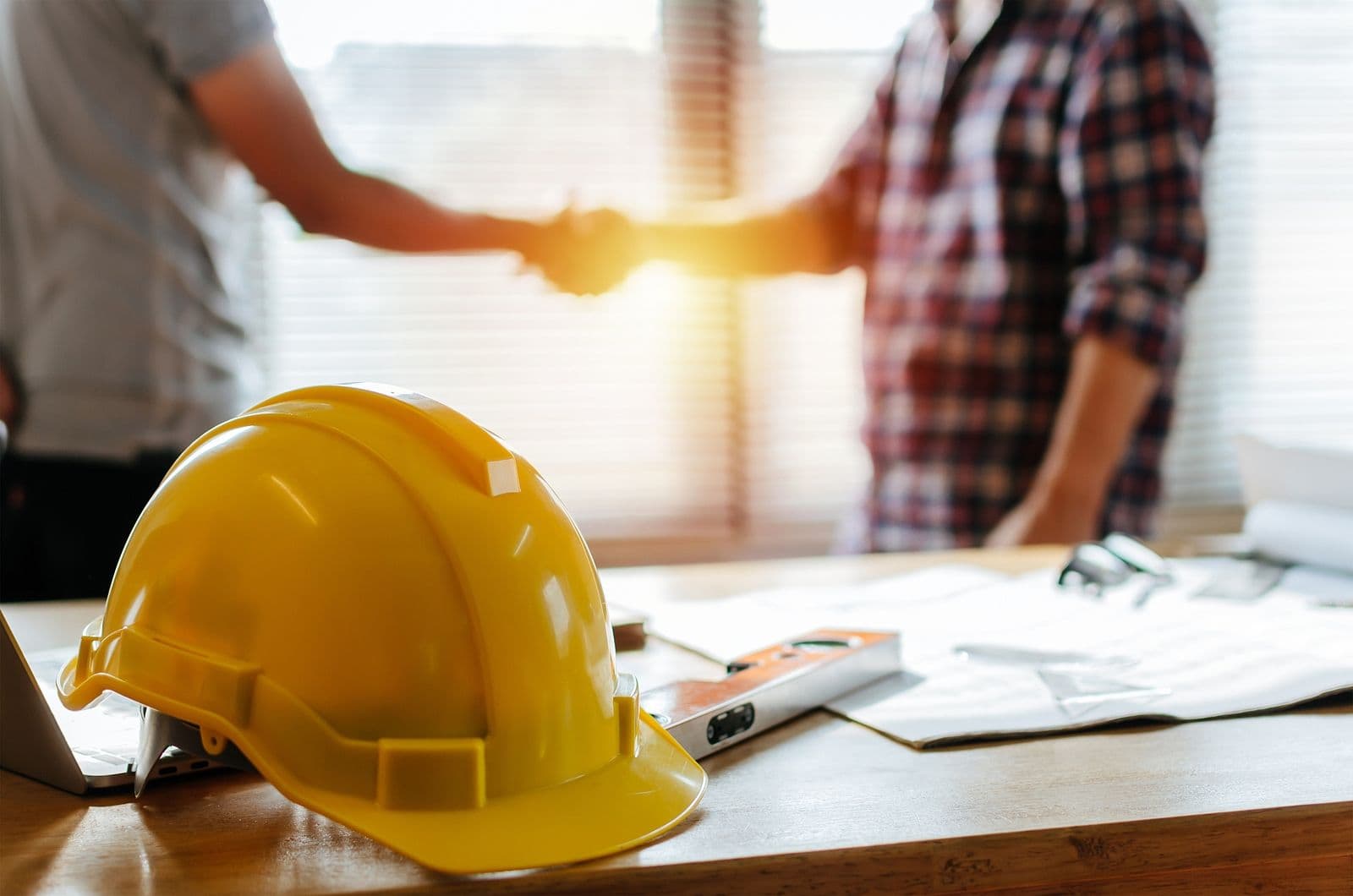 A yellow safety helmet sits on a workbench while people shake hands in the background.