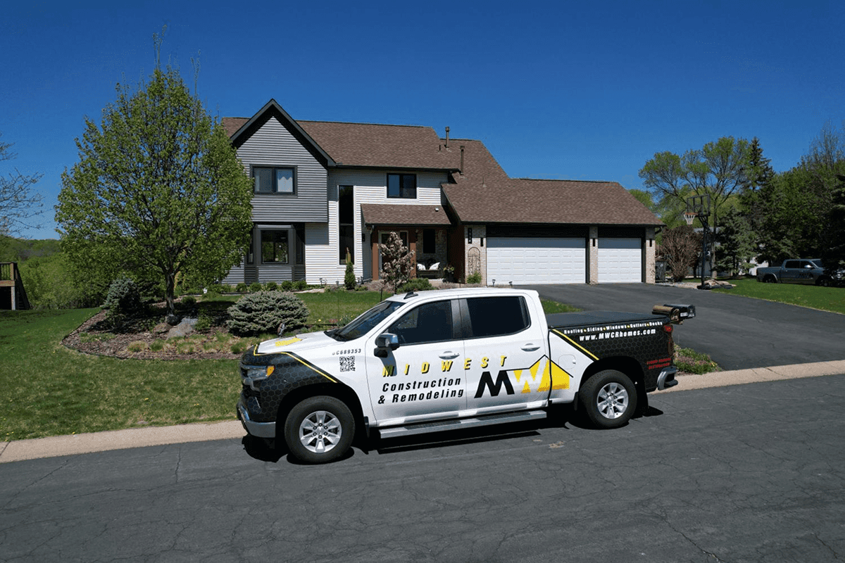 A Midwest Construction & Remodeling truck parked in front of a white and gray house with new brown shingle roofing.
