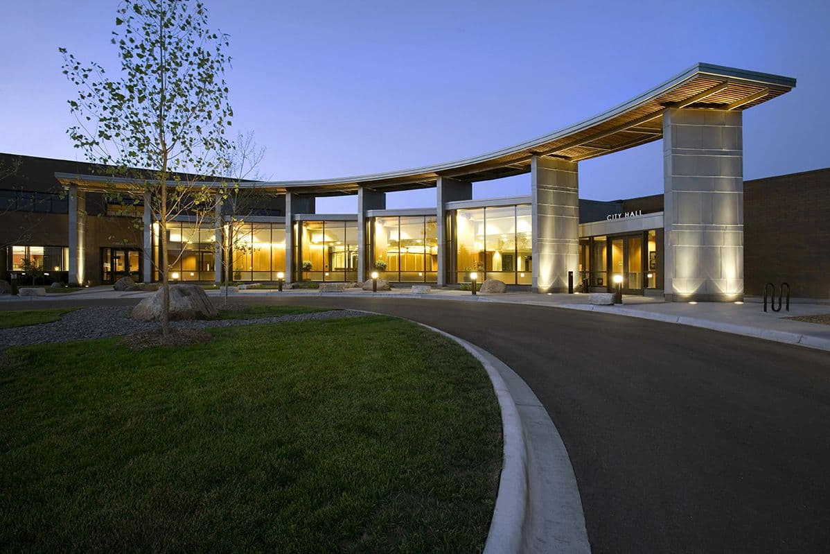 Plymouth City Hall illuminated at dusk with a modern curved roof, glass entrance, landscaped lawn, and driveway in front.