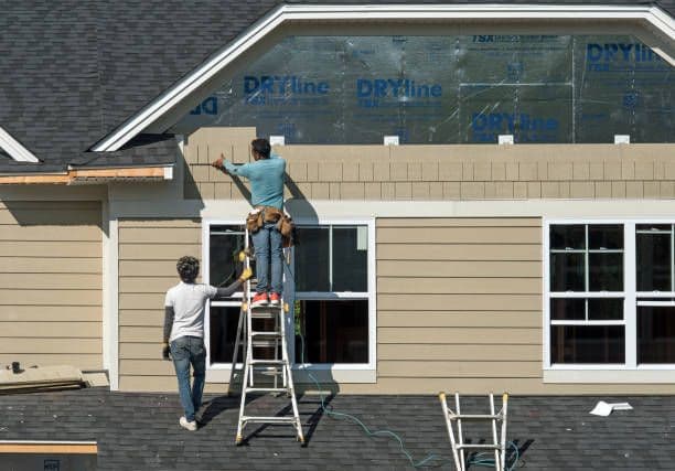 Workers install tan siding on the second story of a house.
